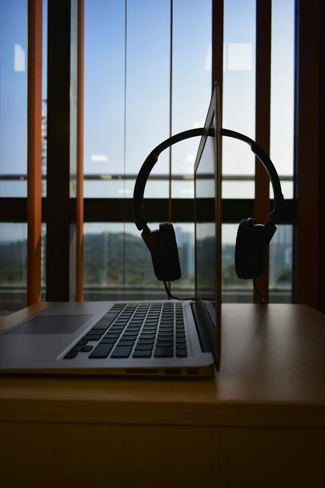 A laptop and headphones on a table beside a window looking out over hills