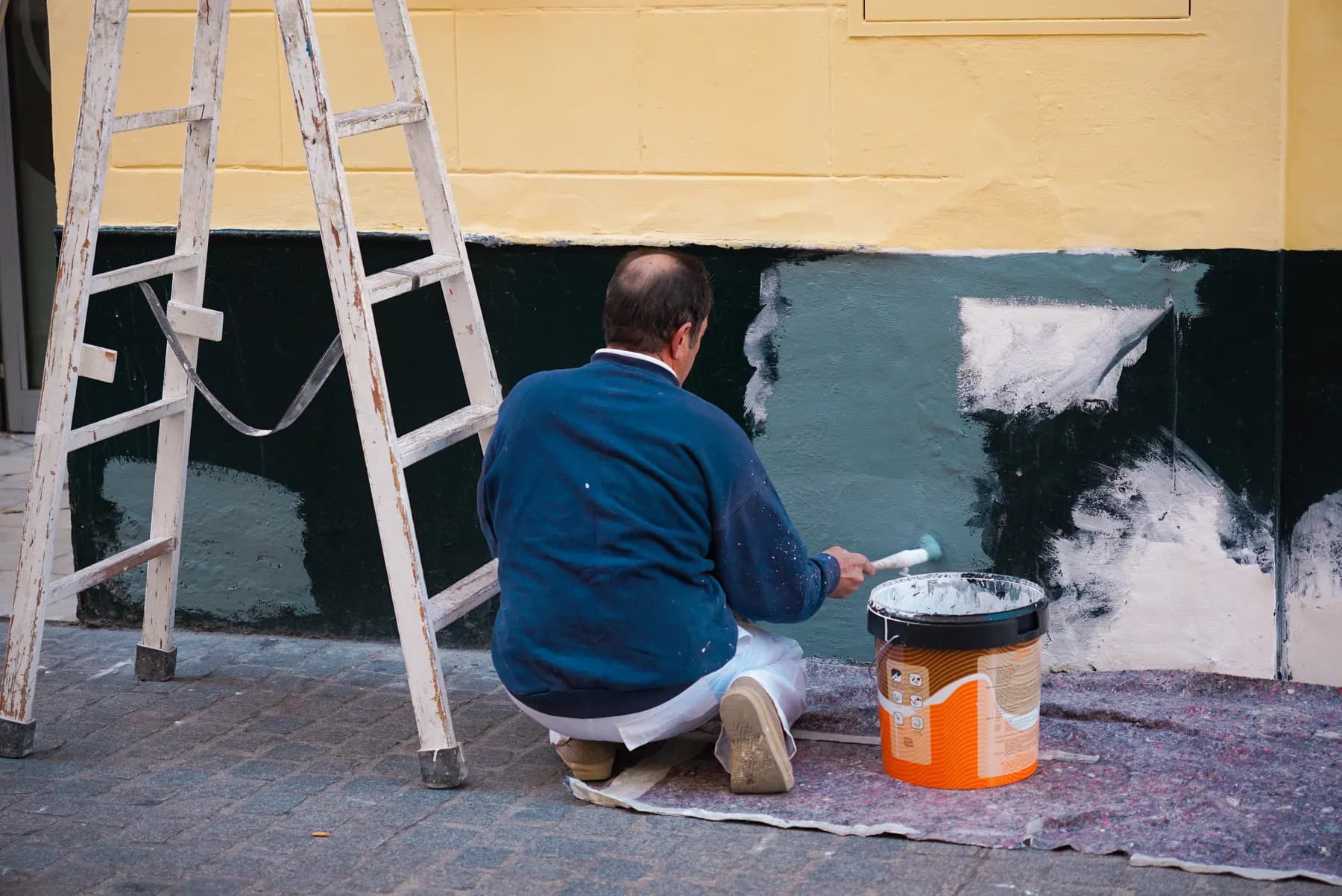 A worker kneeling on a sidewalk beside a paint can, rolling fresh paint onto a wall